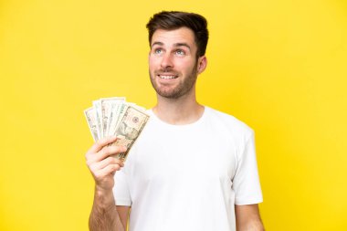 Young caucasian man taking a lot of money isolated on yellow background thinking an idea while looking up