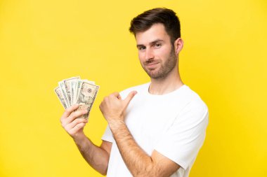 Young caucasian man taking a lot of money isolated on yellow background proud and self-satisfied