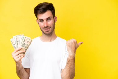Young caucasian man taking a lot of money isolated on yellow background pointing to the side to present a product