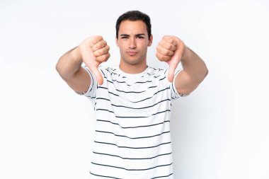 Beyaz arka planda izole edilmiş genç beyaz bir adam baş parmağını iki eliyle aşağı gösteriyor.