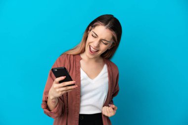 Mavi arka planda izole edilmiş genç beyaz kadın cep telefonu kullanıyor ve zafer jesti yapıyor.
