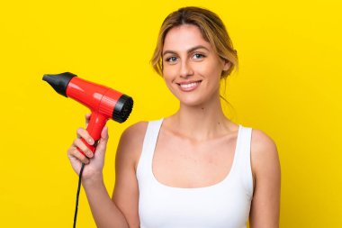 Young Uruguayan woman holding a hairdryer isolate don yellow background smiling a lot