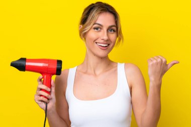 Young Uruguayan woman holding a hairdryer isolate don yellow background pointing to the side to present a product