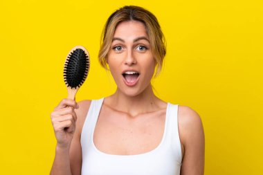 Young Uruguayan woman with hair comb isolated on yellow background with surprise and shocked facial expression