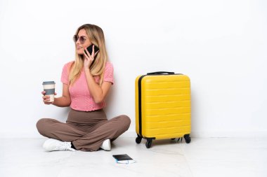Uruguaylı gezgin kadın, beyaz arka planda izole bir şekilde yerde oturuyor. Elinde kahve ve cep telefonu var.