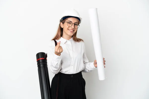 Young architect woman with helmet and holding blueprints isolated on white background making money gesture