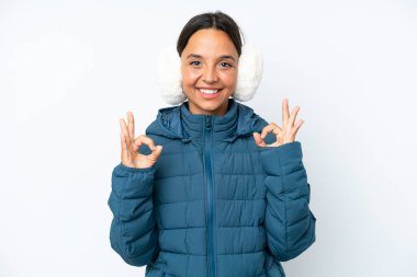 Young hispanic woman wearing a winter earmuffs isolated on white background showing an ok sign with fingers
