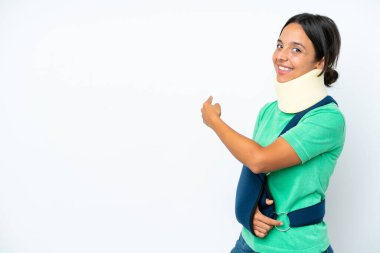 Young hispanic woman wearing a neck brace and sling isolated on white background pointing back