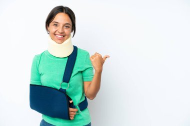 Young hispanic woman wearing a neck brace and sling isolated on white background pointing to the side to present a product