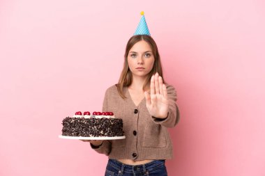 Young Lithuanian woman holding birthday cake isolated on pink background making stop gesture