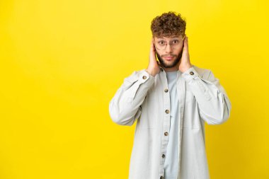 Sarı arka planda izole edilmiş yakışıklı beyaz adam hayal kırıklığına uğramış ve kulaklarını kapatmış.