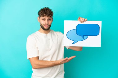 Genç beyaz adam mavi arka planda izole edilmiş. Elinde konuşma balonu ikonu olan bir levha var.