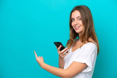 Genç beyaz kadın mavi arka planda izole edilmiş cep telefonu kullanıyor ve geri işaret ediyor.