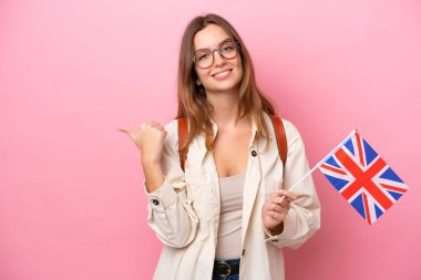 Beyaz Amerikalı genç bir kadın pembe arka planda izole edilmiş bir Birleşik Krallık bayrağı tutuyor ve bir ürünü sunmak için kenarı işaret ediyor.