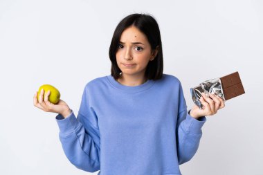 Beyaz arka planda izole olmuş genç beyaz bir kadın bir elinde çikolata tableti, diğer elinde elma tutarken kuşkuları var.