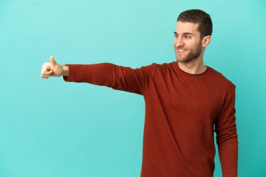 Yakışıklı sarışın, soyutlanmış mavi arka planda, başparmak işareti yapıyor.