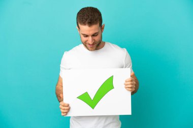 İzole edilmiş mavi arkaplanı olan yakışıklı sarışın adam. Üzerinde yeşil işaret simgesi olan bir pankart tutuyor.