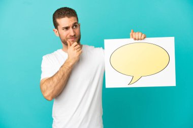 Yakışıklı sarışın, izole edilmiş mavi arkaplan üzerinde konuşma baloncuğu ikonu olan bir levha tutuyor ve düşünüyor.