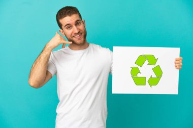 Yakışıklı sarışın adam, izole edilmiş mavi arka planda elinde geri dönüşüm simgesi olan bir pankart tutuyor ve telefon hareketi yapıyor.