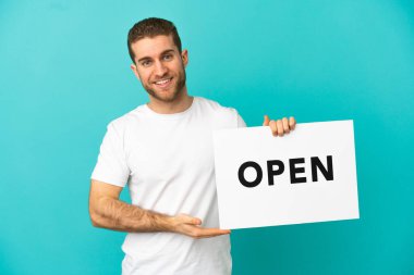 Yakışıklı, mavi arka planda, üzerinde mutlu bir ifade olan barış sembolü olan bir pankart tutan sarışın adam.