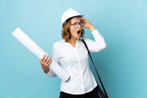 Young architect Georgian woman with helmet and holding blueprints over isolated background doing surprise gesture while looking to the side