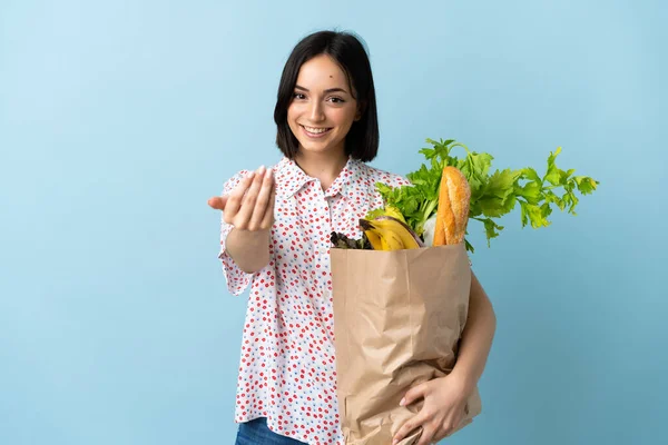 Young woman holding a grocery shopping bag inviting to come with hand. Happy that you came