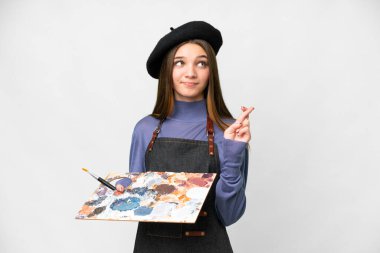 Young artist man holding a palette over isolated white background with fingers crossing and wishing the best