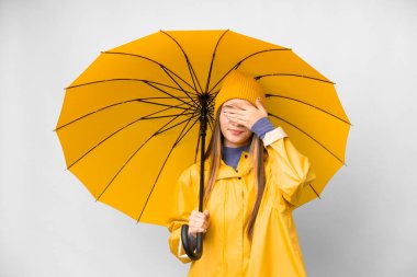 Teenager girl with rainproof coat and umbrella over isolated white background covering eyes by hands. Do not want to see something