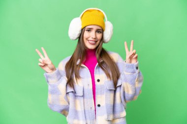 Young woman wearing winter muffs over isolated chroma key background showing victory sign with both hands