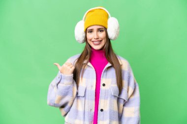 Young woman wearing winter muffs over isolated chroma key background pointing to the side to present a product