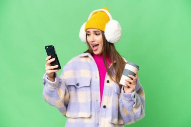 Young woman wearing winter muffs over isolated chroma key background holding coffee to take away and a mobile
