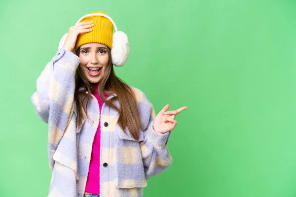 Young woman wearing winter muffs over isolated chroma key background surprised and pointing finger to the side