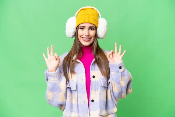 Young woman wearing winter muffs over isolated chroma key background showing an ok sign with fingers