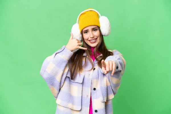 Young woman wearing winter muffs over isolated chroma key background making phone gesture and pointing front