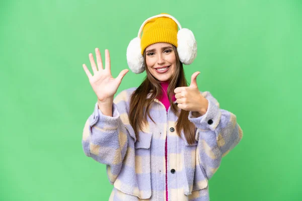 Young woman wearing winter muffs over isolated chroma key background counting six with fingers