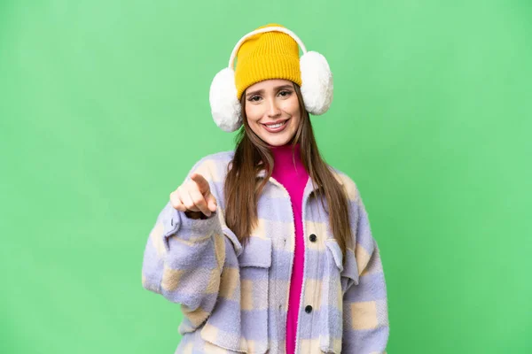 Young woman wearing winter muffs over isolated chroma key background points finger at you with a confident expression