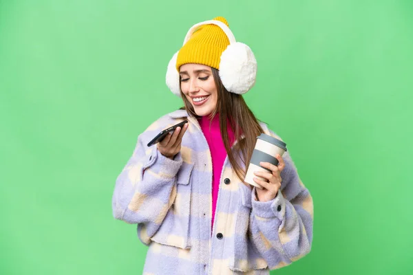 Young woman wearing winter muffs over isolated chroma key background holding coffee to take away and a mobile
