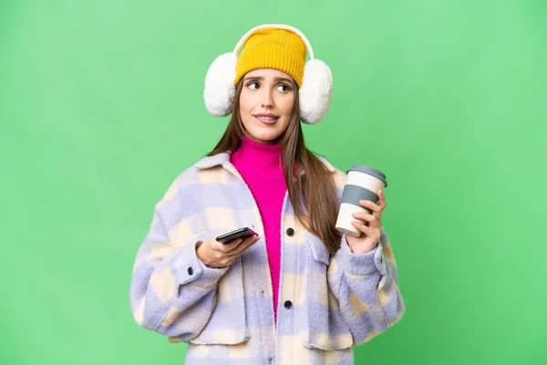 Young woman wearing winter muffs over isolated chroma key background holding coffee to take away and a mobile while thinking something