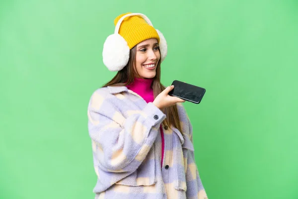 Young woman wearing winter muffs over isolated chroma key background keeping a conversation with the mobile phone with someone