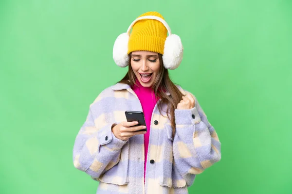 Young woman wearing winter muffs over isolated chroma key background surprised and sending a message