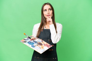 Young artist woman holding a palette over isolated chroma key background and looking up