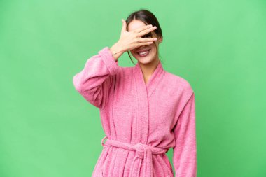 Young beautiful woman in a bathrobe over isolated chroma key background covering eyes by hands and smiling