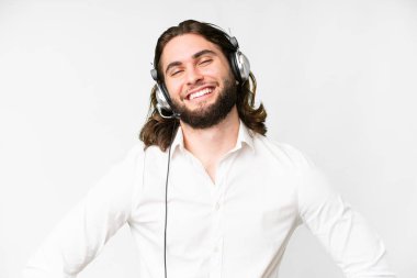 Telemarketer man working with a headset over isolated white background posing with arms at hip and smiling