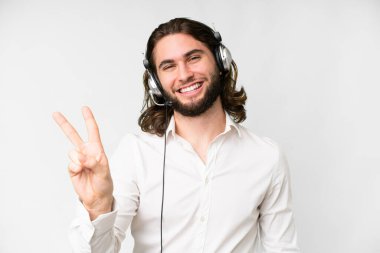Telemarketer man working with a headset over isolated white background smiling and showing victory sign