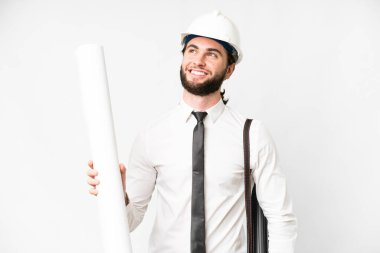 Young architect man with helmet and holding blueprints over isolated white background thinking an idea while looking up