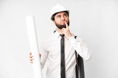 Young architect man with helmet and holding blueprints over isolated white background having doubts while looking up