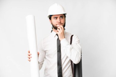 Young architect man with helmet and holding blueprints over isolated white background having doubts and thinking