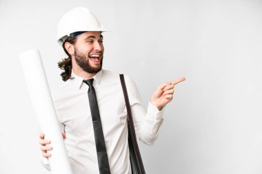 Young architect man with helmet and holding blueprints over isolated white background pointing finger to the side and presenting a product
