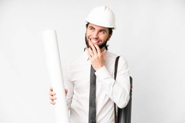 Young architect man with helmet and holding blueprints over isolated white background looking up while smiling