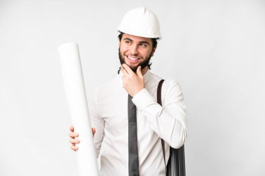 Young architect man with helmet and holding blueprints over isolated white background looking to the side and smiling